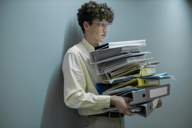 An overworked office worker carrying a large stack of files and folders in a modern setting.
